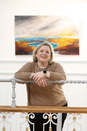 Person in brown sweater and black pants leaning on white decorative railing at sunset with orange sky in background.