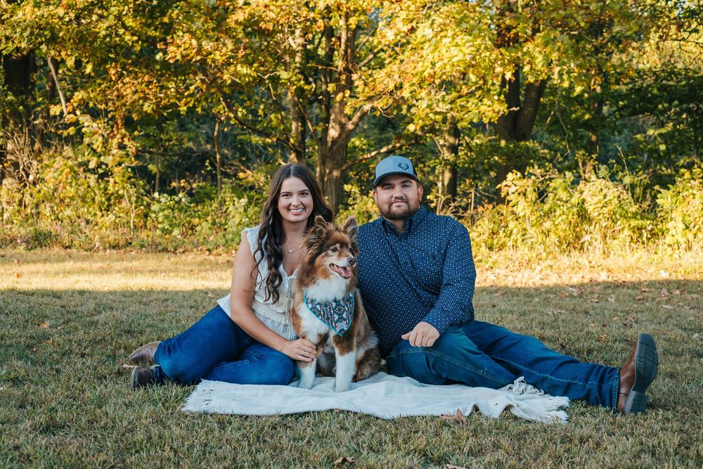 A couple relaxes on a white blanket in a sunlit autumn field during a casual outdoor photo session.