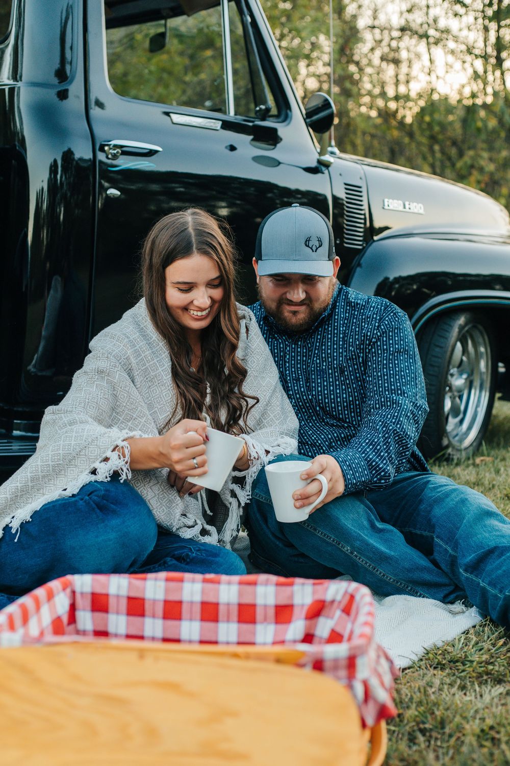 A couple shares coffee while sitting on a picnic blanket next to a vintage black truck during sunset.