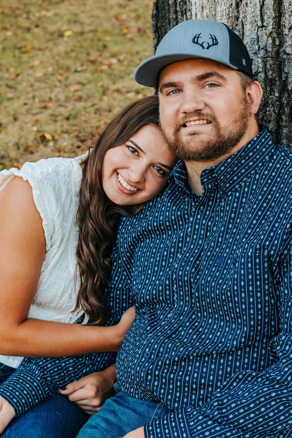 A couple in casual attire poses together against a tree trunk in an autumn setting.