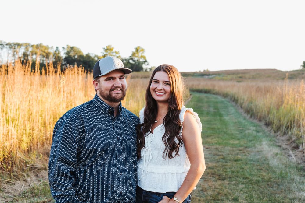 A couple poses together in a golden field during sunset while capturing photos.