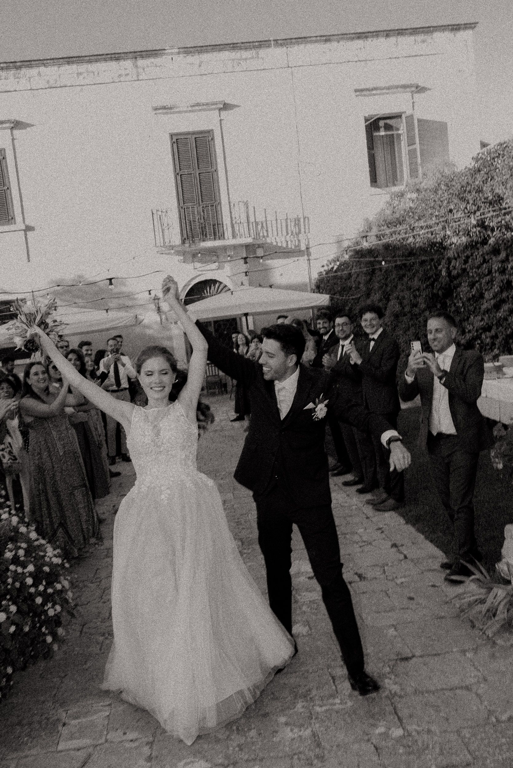 A sequence of black and white wedding photos showing a couple celebrating and dancing with guests outside a building.