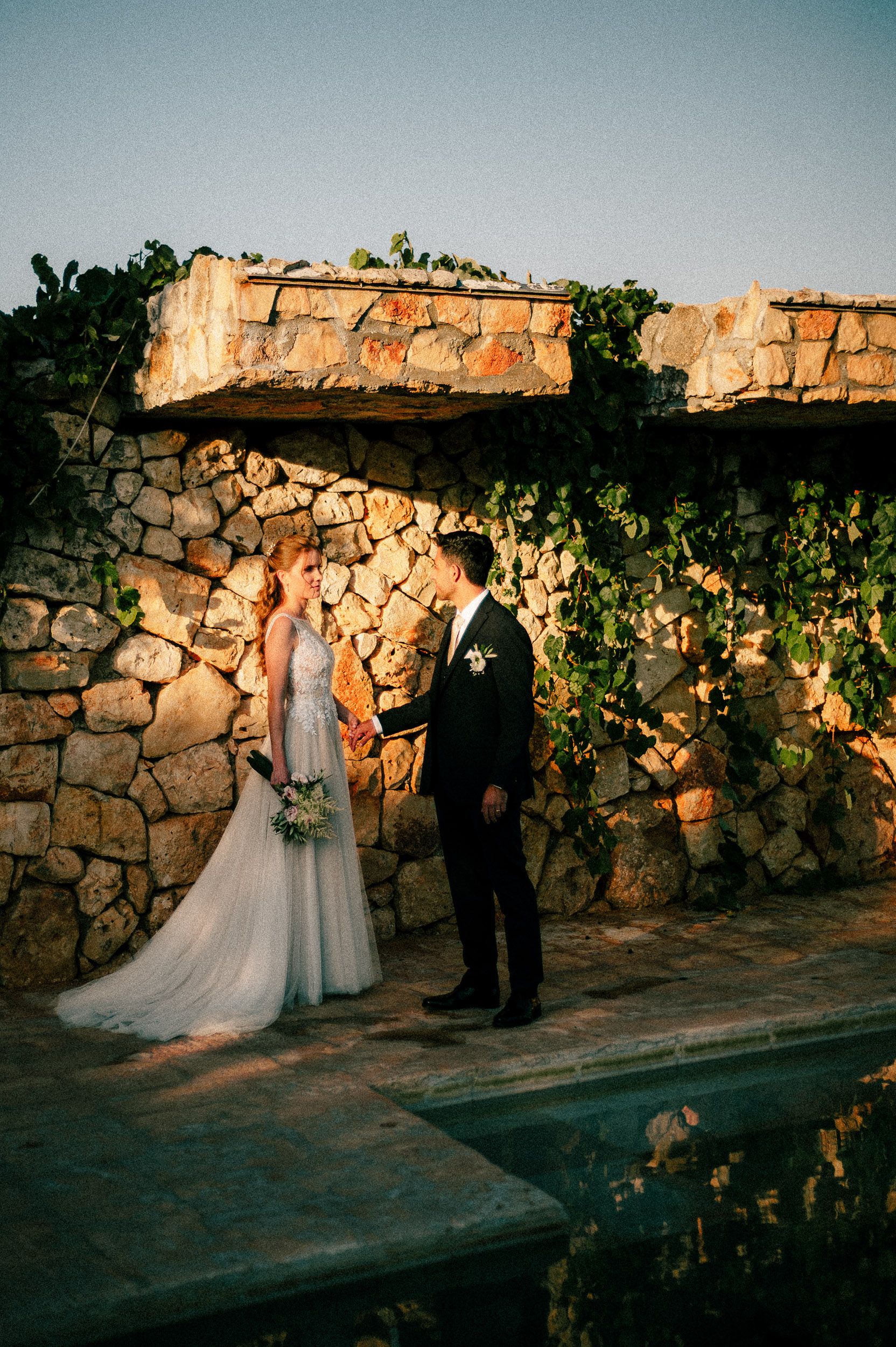 A couple stands by a rustic stone wall at sunset during their wedding ceremony at an outdoor venue.