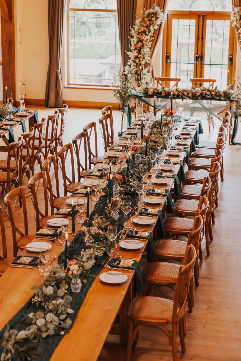 Long wooden dining tables set for a wedding reception with elegant place settings and eucalyptus garland centerpieces.