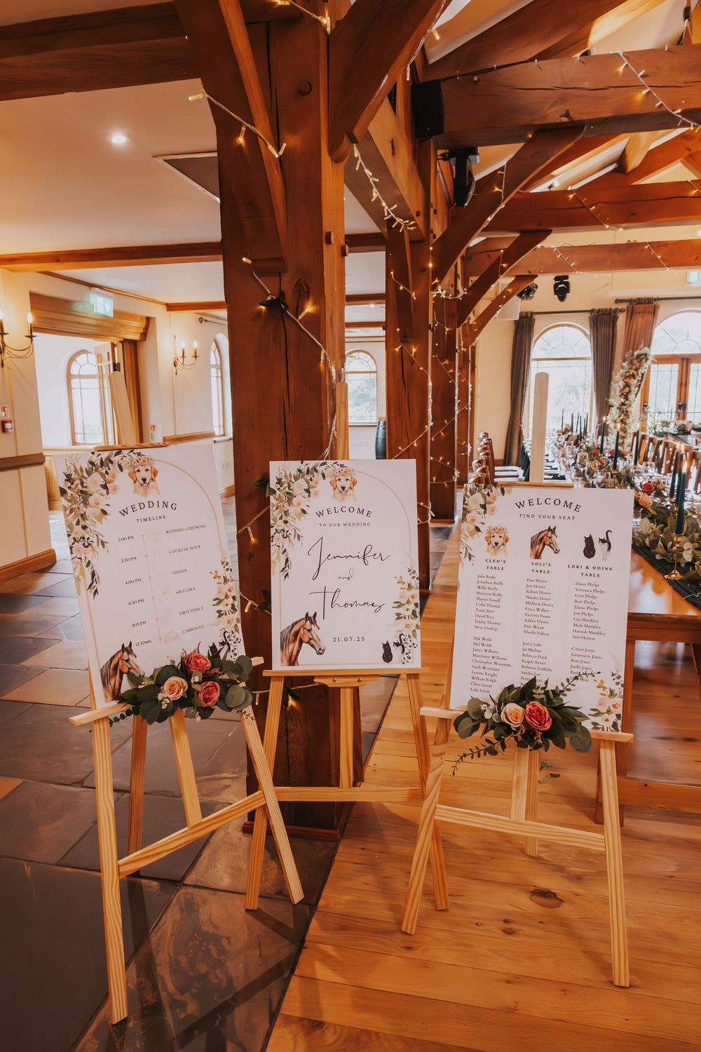 Wedding seating chart displayed on wooden easels with floral decorations in a rustic venue with exposed wooden beams.