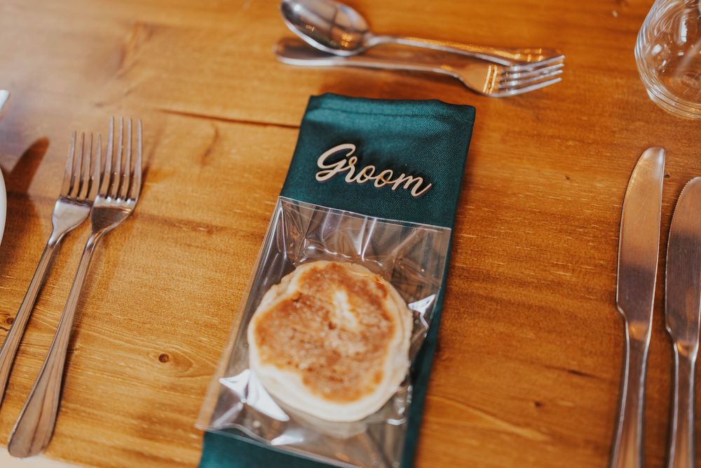 A place setting at a wooden table with silverware and a green napkin containing a round baked pastry.