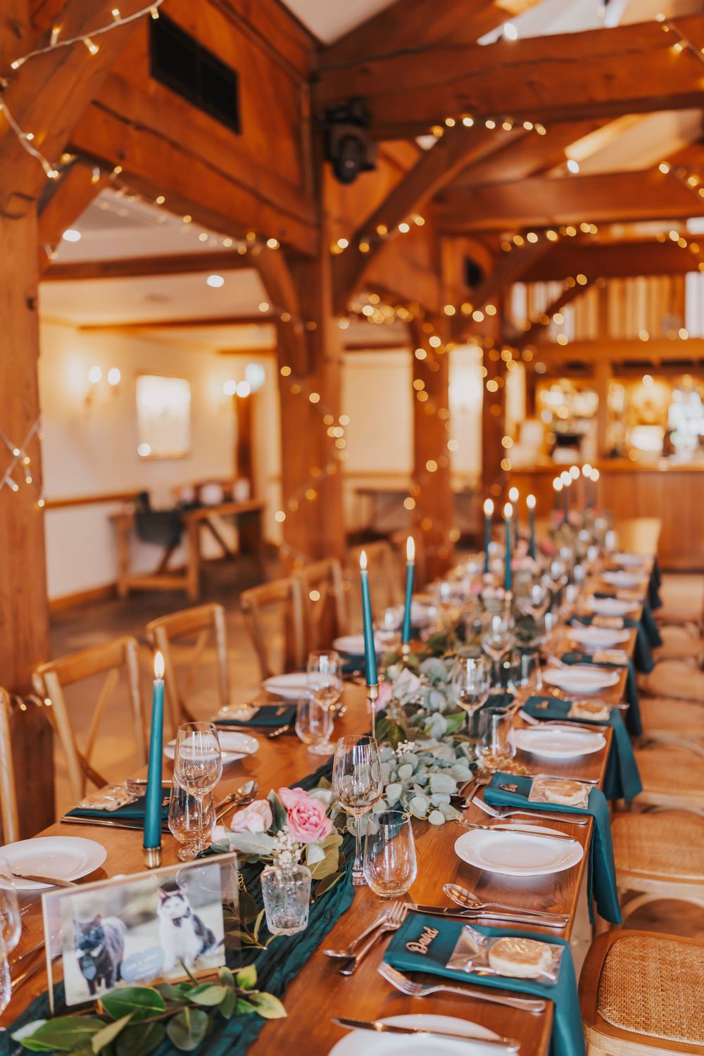 Elegant wedding reception table setup with teal napkins, string lights, and wooden beams in rustic barn venue.