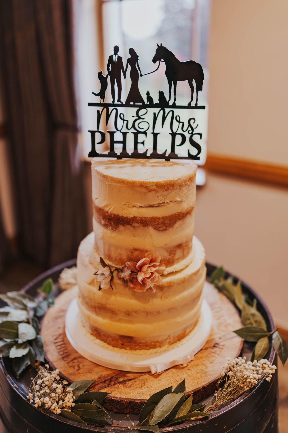 A rustic semi-naked wedding cake decorated with greenery and silhouette cake toppers on a white plate.