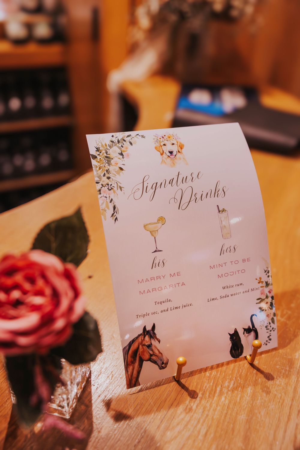 Elegant wedding signature drink menu cards with floral designs and cat illustrations displayed on wooden surfaces.