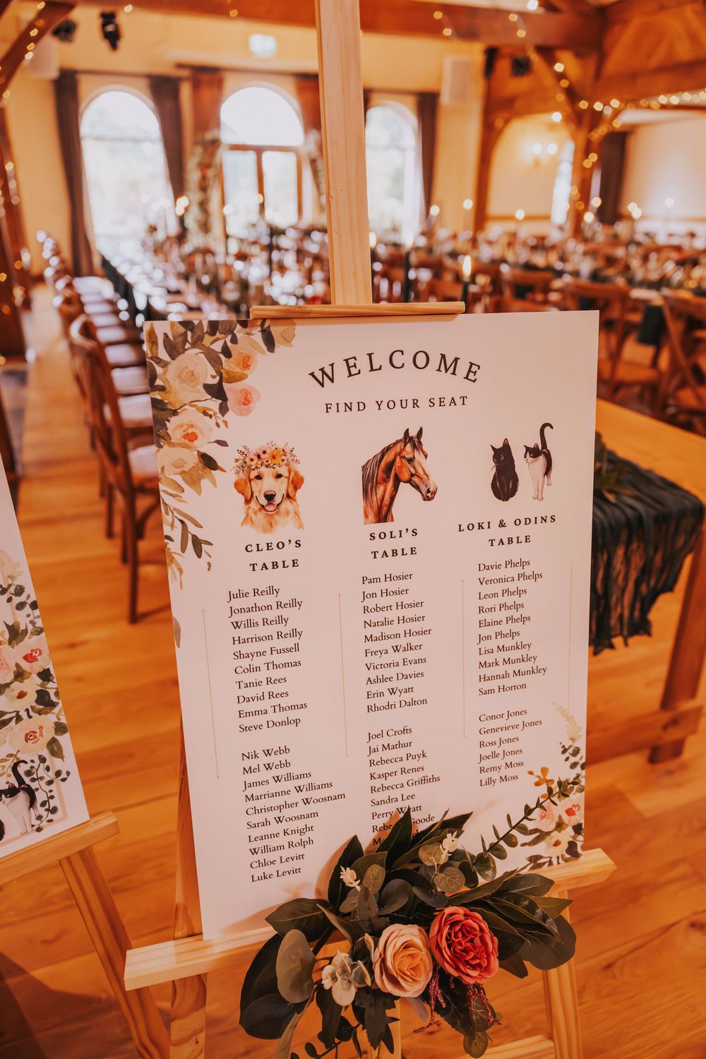 Wedding reception seating chart displays with floral decorations and table assignments on elegant white cards at a warm-lit venue.