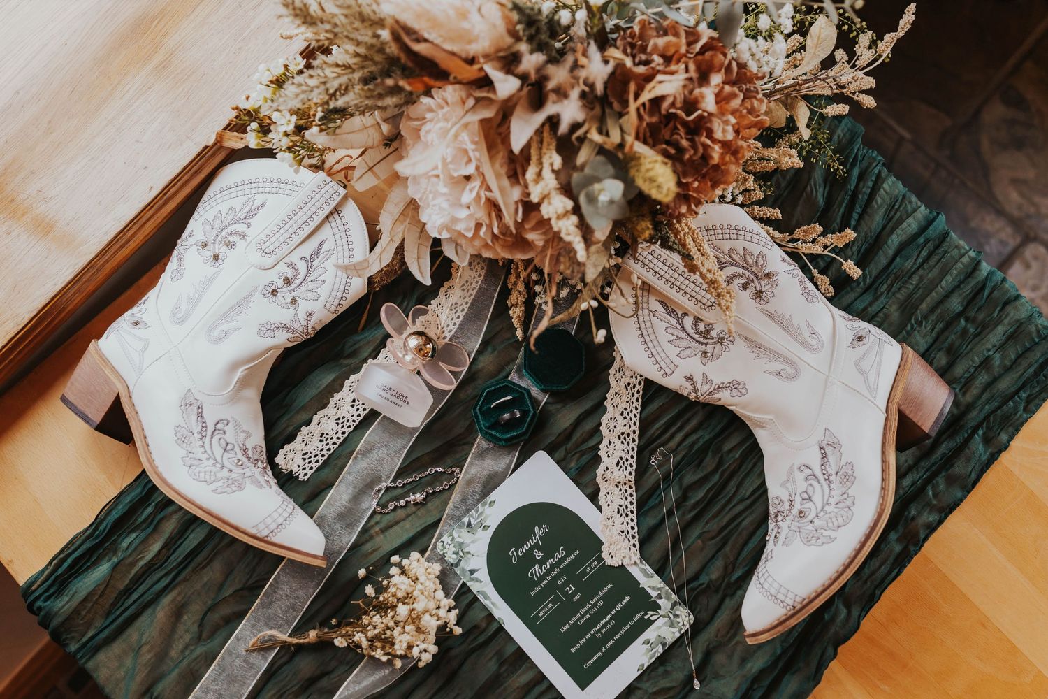 White leather cowboy boots with floral embroidery displayed next to dried flowers on a green velvet surface.