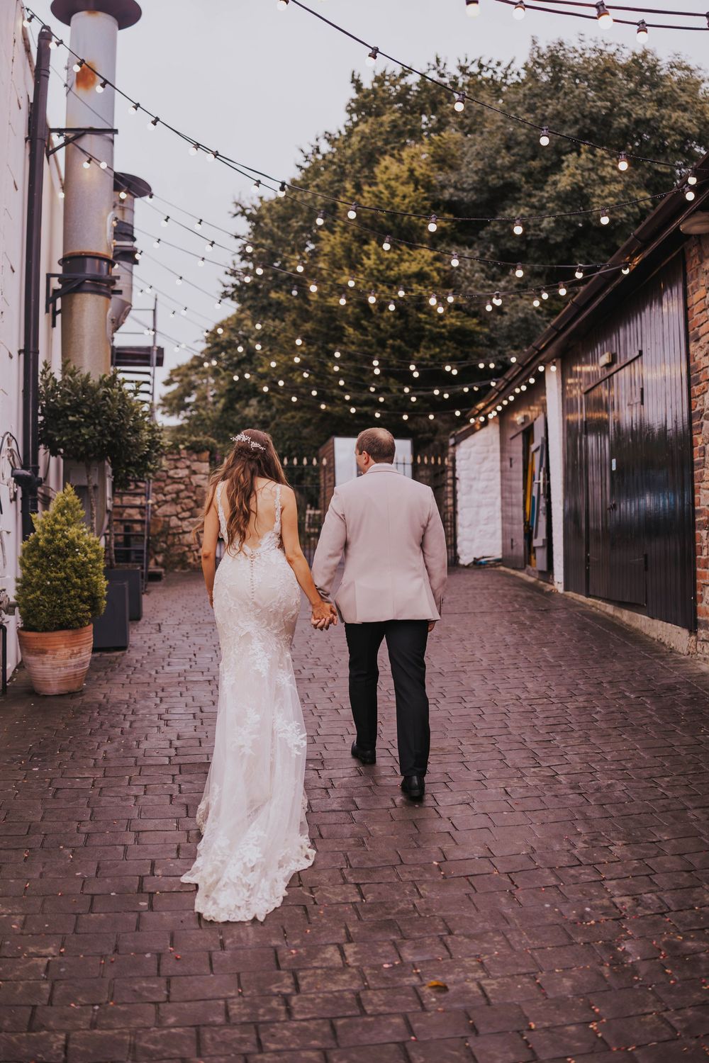 A romantic couple walks down a cobblestone path under twinkling string lights at dusk.