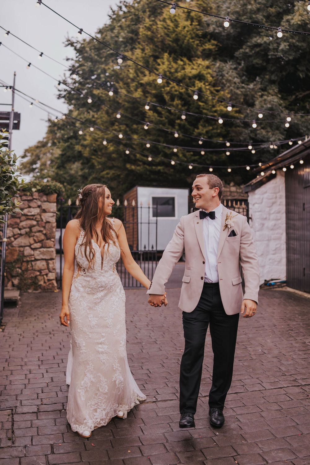 Couple walking hand in hand on brick pathway under string lights at outdoor wedding venue during sunset.