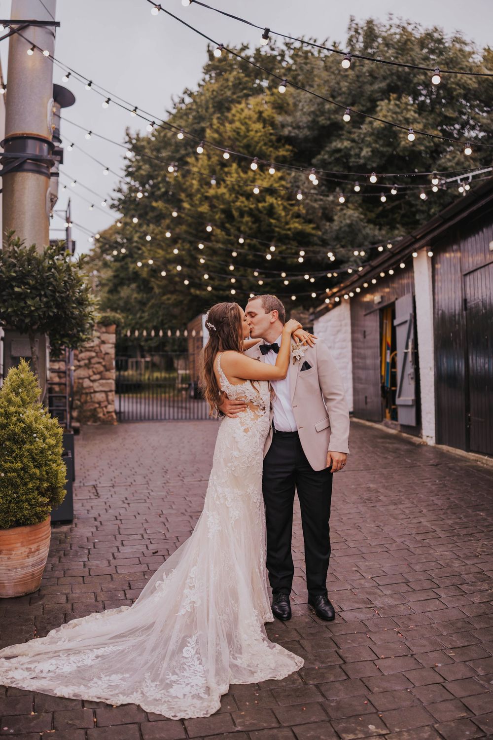 A couple shares a romantic moment under string lights at an outdoor wedding venue with brick paving and greenery.