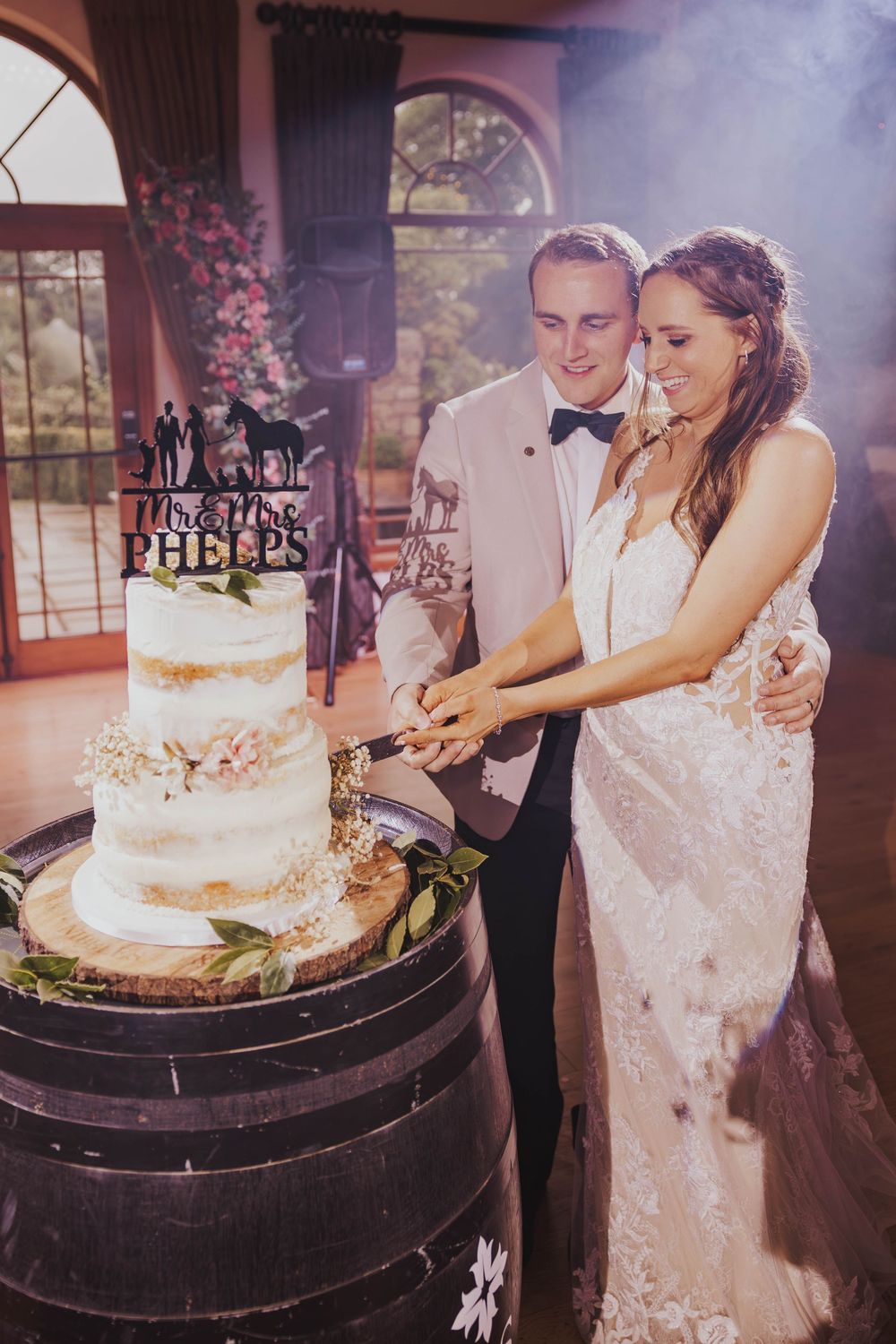 A couple cuts their elegant white semi-naked wedding cake displayed on a rustic wine barrel at an indoor venue.