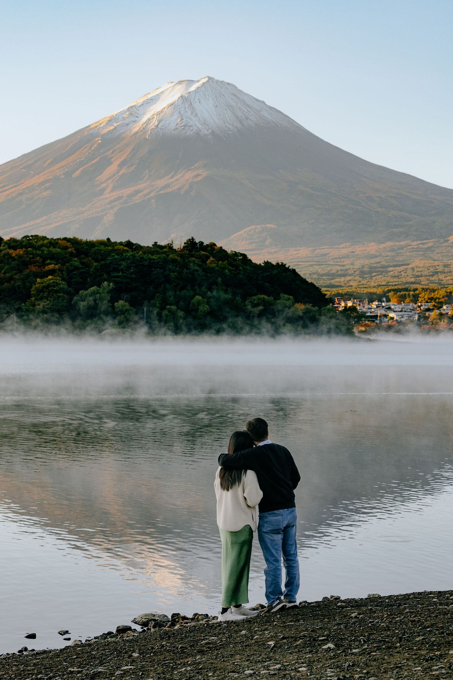 Two figures stand silhouetted by the misty shores of Lake Yamanaka with Mount Fuji rising majestically in the background.
