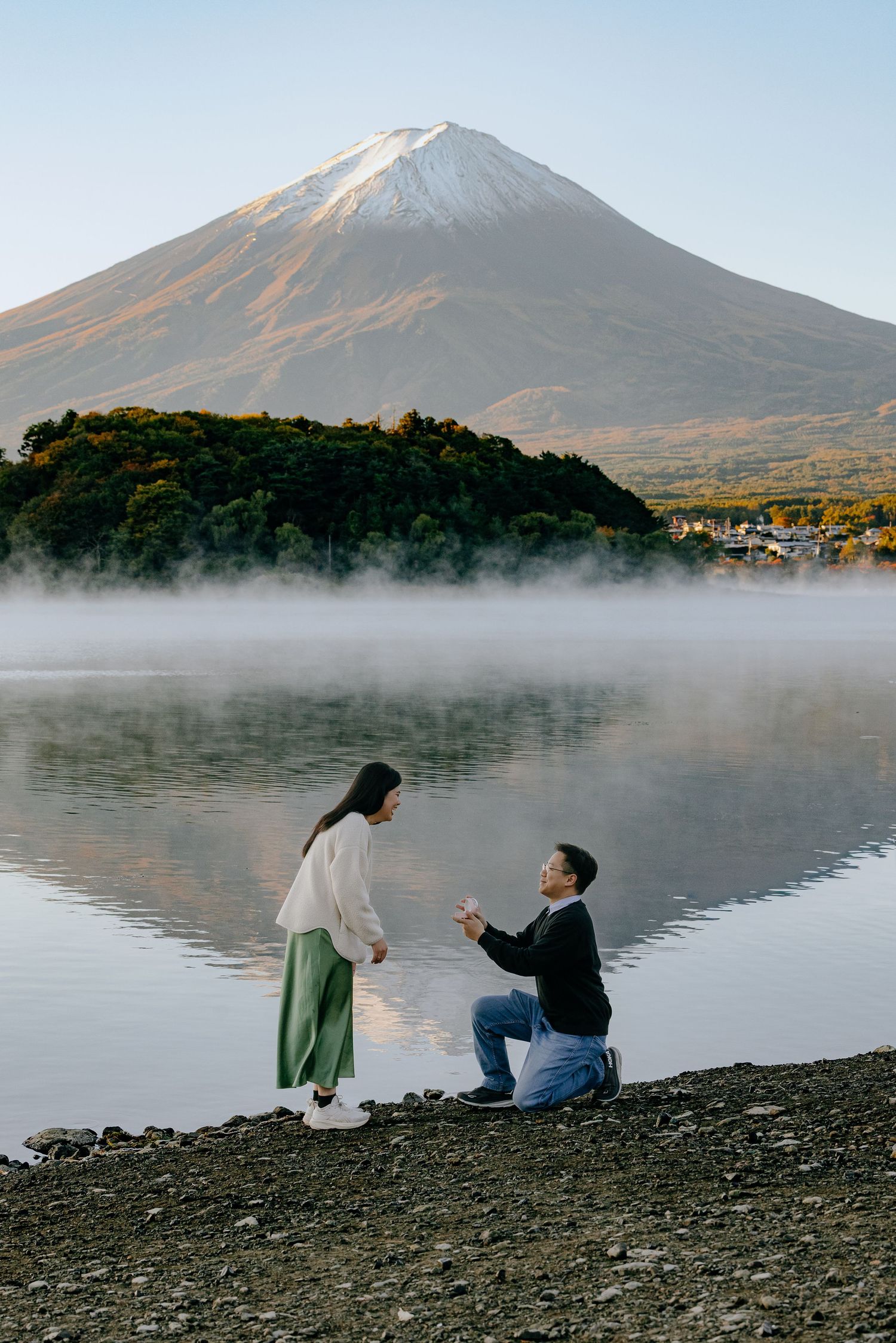 Two people sit on a rocky shore by Lake Yamanaka with Mount Fuji rising majestically in the misty background.