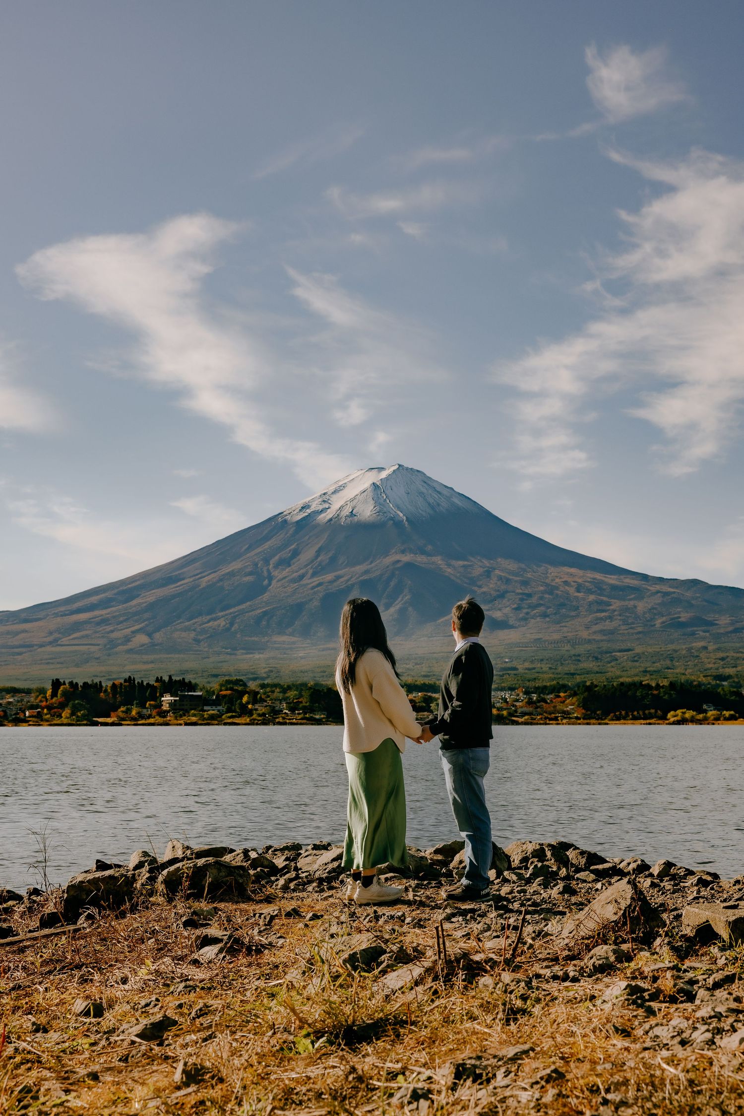 Mount Fuji rises majestically over Lake Kawaguchiko as two figures admire the iconic Japanese landscape from the shore.