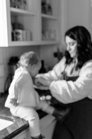 A tender black and white moment between parent and child in a kitchen, with cabinets and shelves visible in the background.