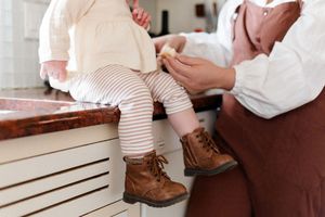 Small brown leather lace-up boots being put on tiny feet wearing striped leggings on wooden storage bench.