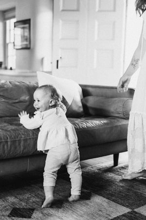Black and white photo of a baby in a white dress and striped pants standing by a leather couch in a living room.