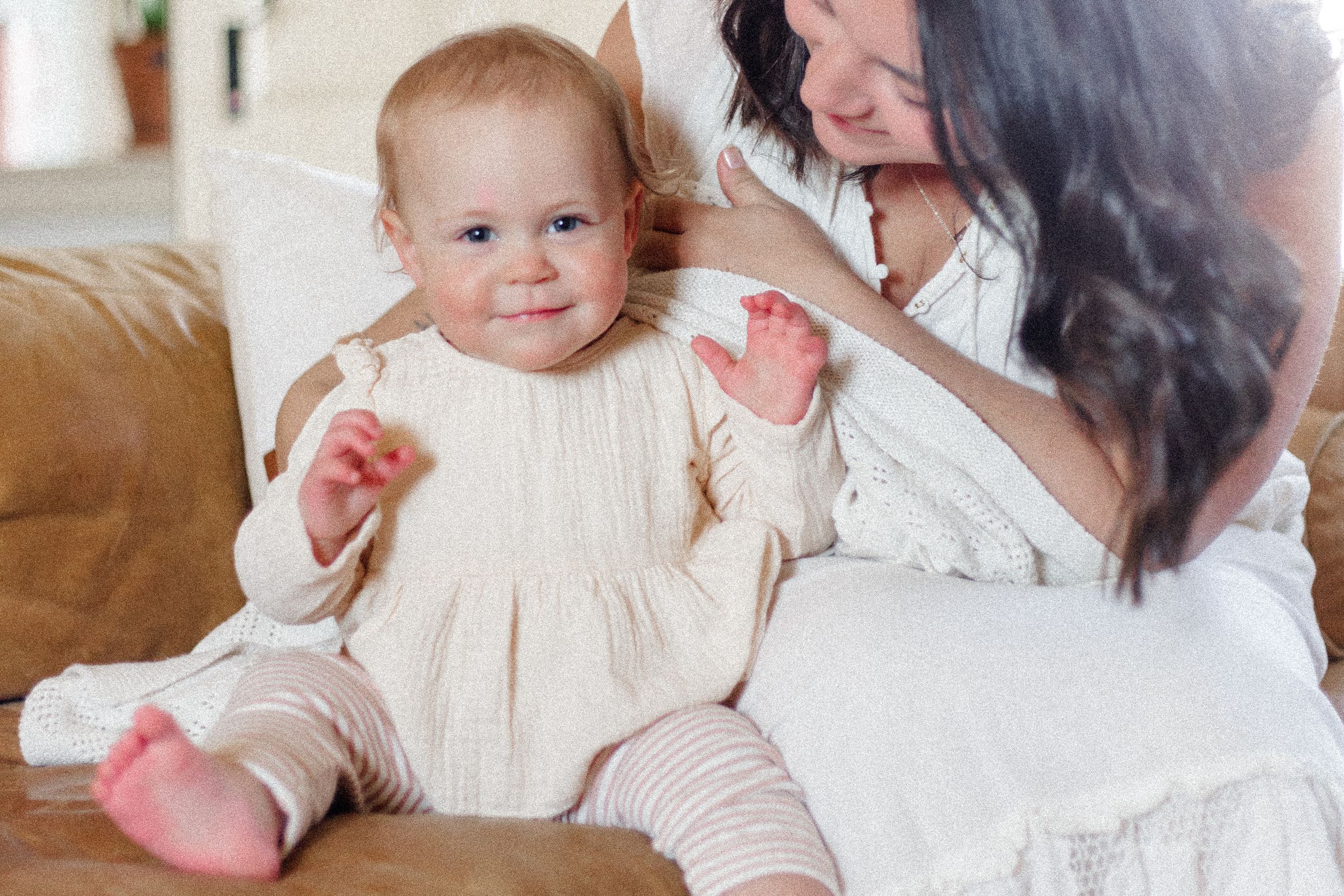 Baby in cream-colored dress sits on brown leather couch with caregiver in white top.