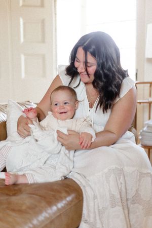A joyful moment shared between parent and baby wearing white outfits while sitting on a brown leather couch.