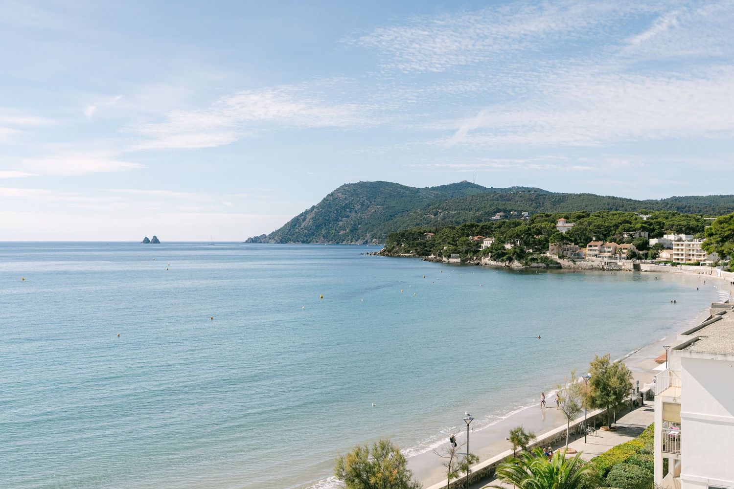 Scenic Mediterranean coastline with turquoise waters, a curved beach, and green hills dotted with coastal buildings.