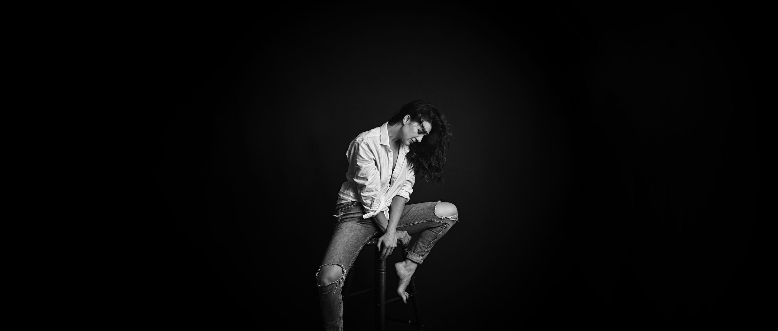 A woman is sitting on a stool, she is wearing a white shirt and ripped jeans. the Image is in black and white.