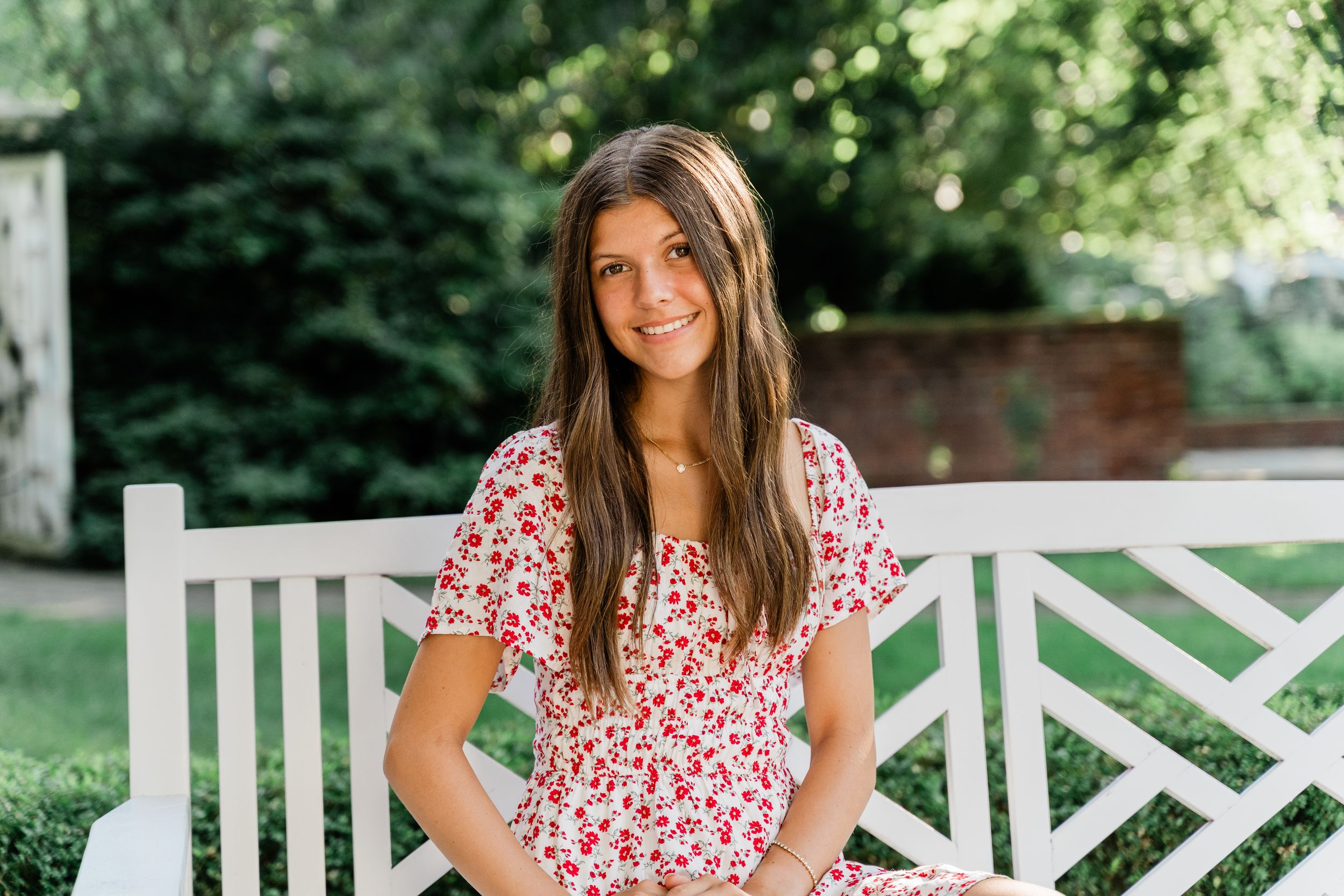 Person in a red and white floral dress sits on a white wooden bench in a sunny garden setting.