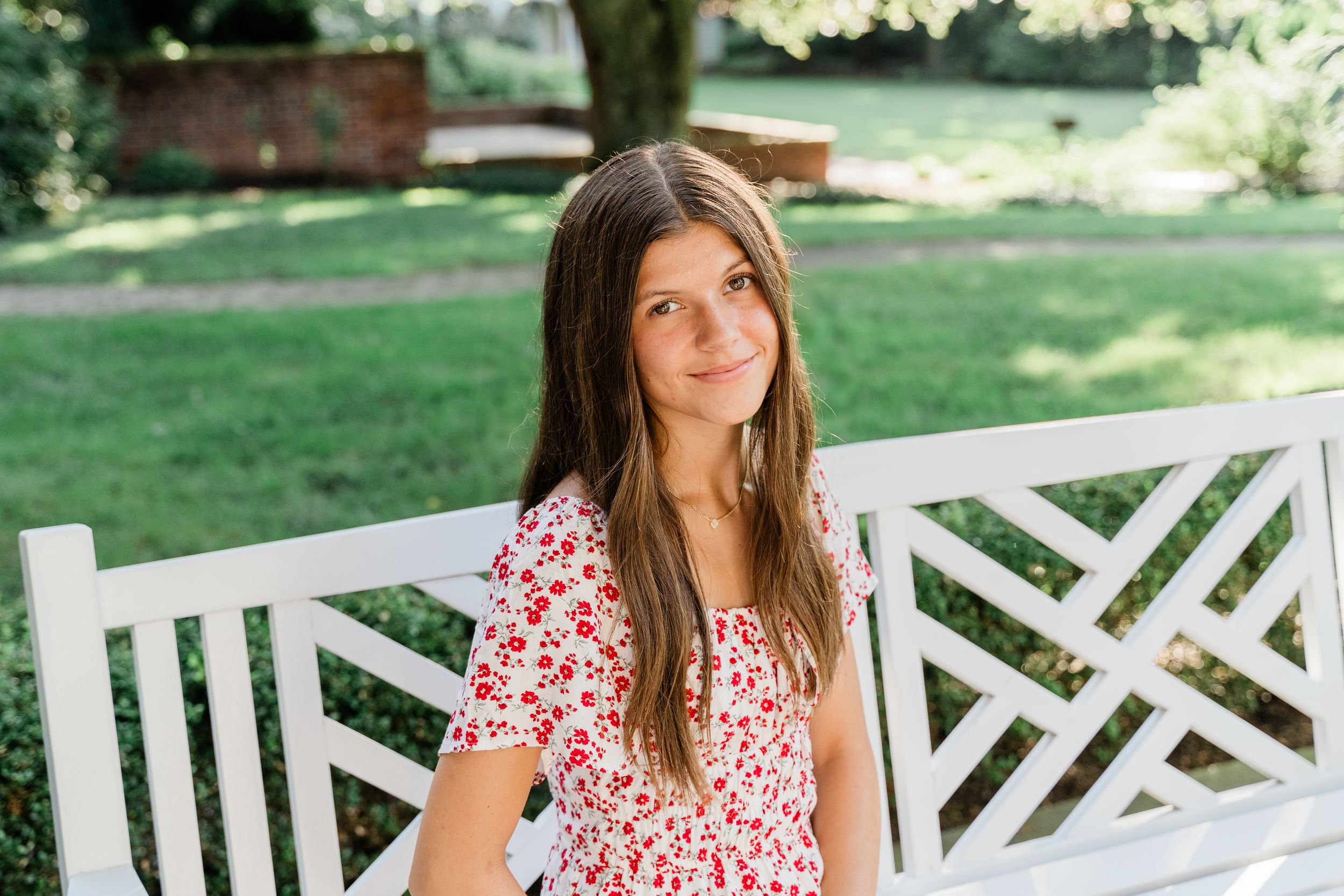 A person in a floral dress poses on a white wooden bench in a sunny garden setting.