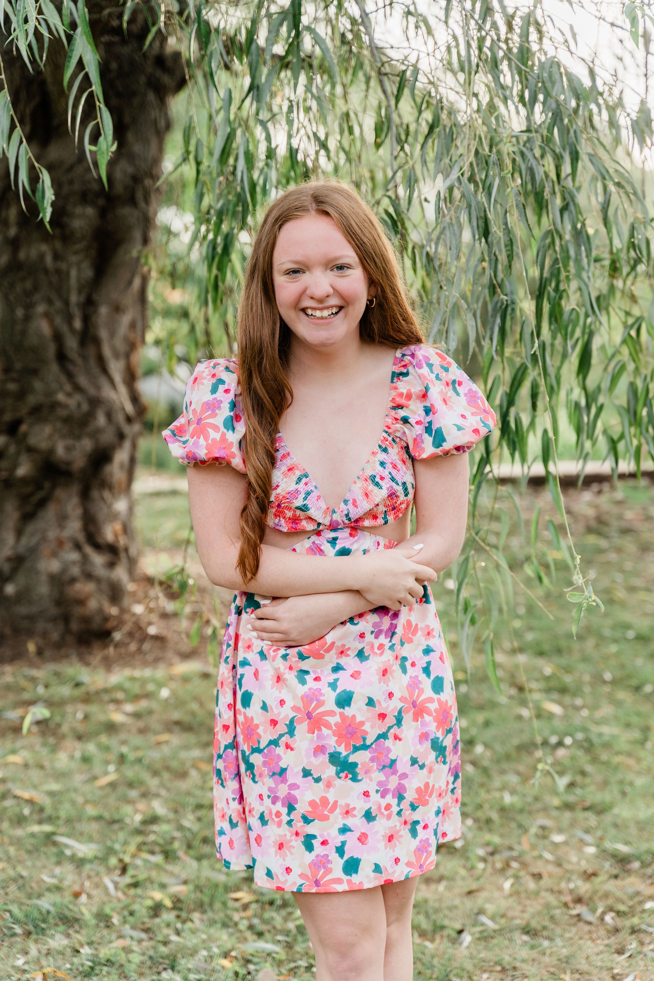 Someone wearing a floral pink and white dress poses outdoors near drooping willow tree branches.