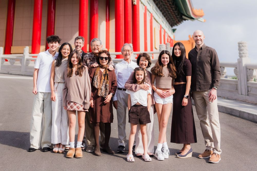 A group stands together smiling outside a traditional Asian building with red pillars on a sunny day.
