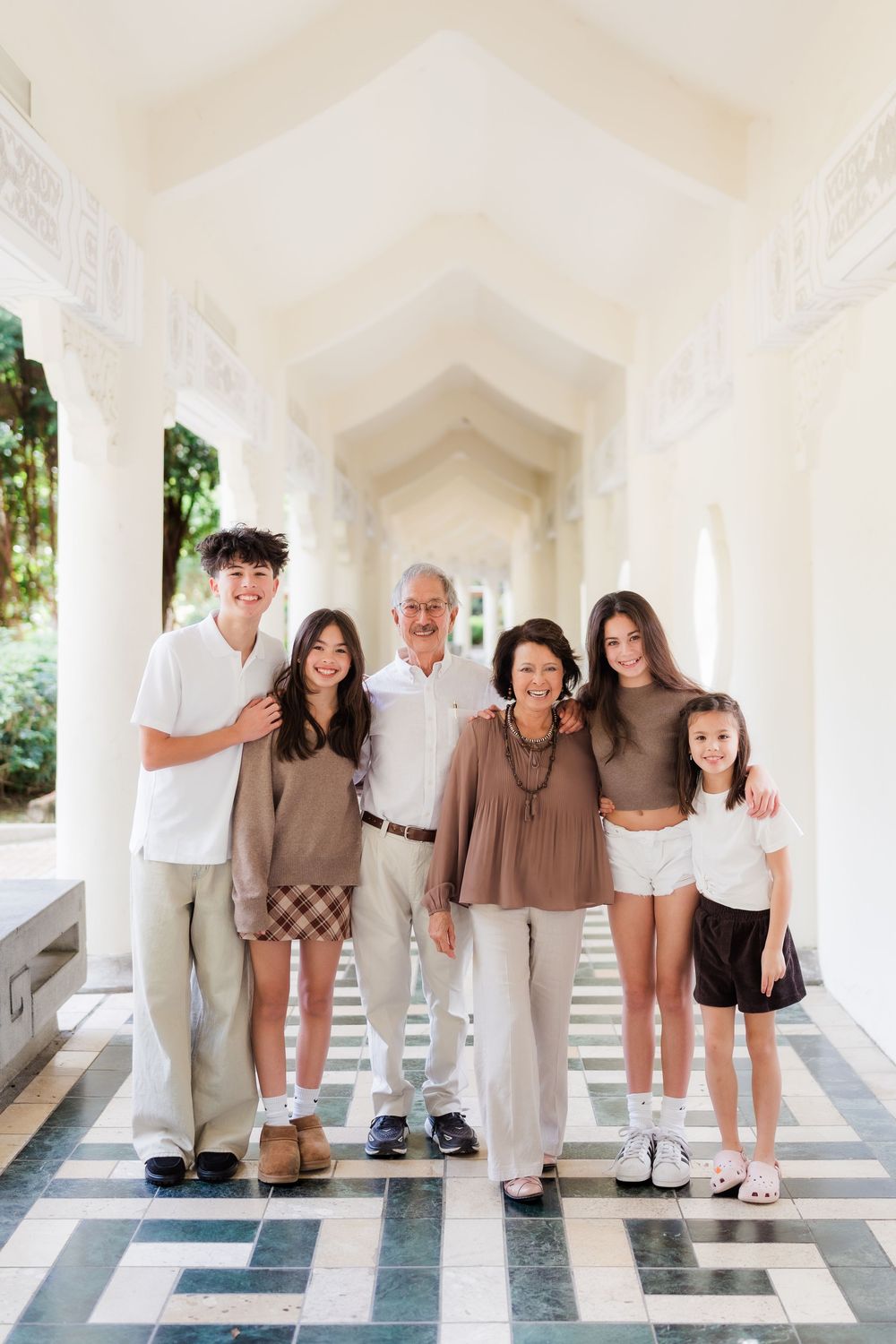 A multi-generational family poses together in a white arched corridor wearing coordinated brown and white outfits.