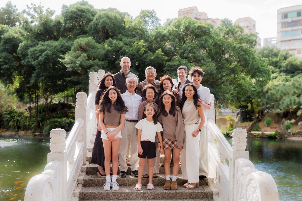 Group photo of wedding party in white and beige dresses posing by a scenic lake surrounded by greenery.