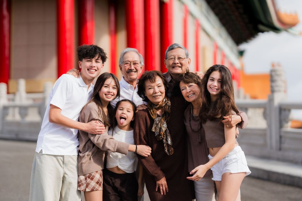 Three generations of family members pose together smiling outside a traditional Asian temple with red pillars.