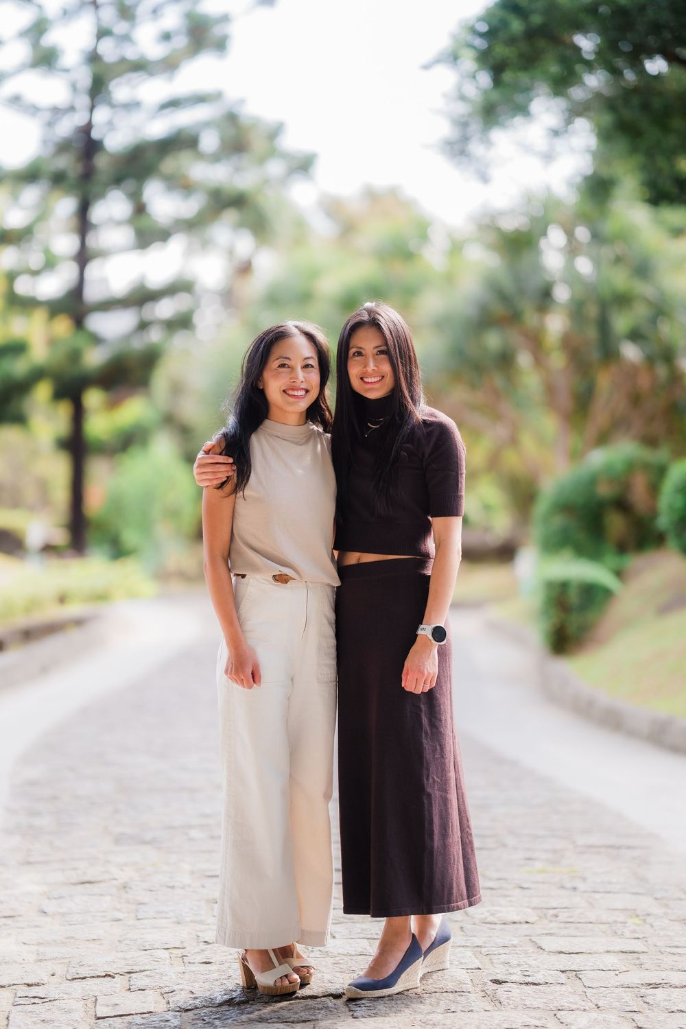Two women in contrasting black and white dresses posing together on a stone path surrounded by trees.