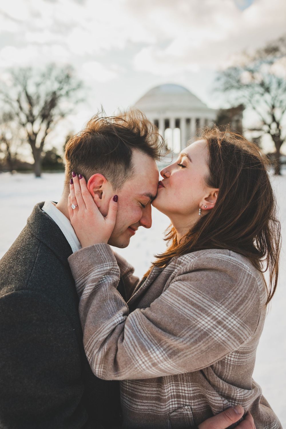 A romantic winter scene shows a couple embracing and kissing while wearing cozy sweaters outdoors in front the jefferson memorial