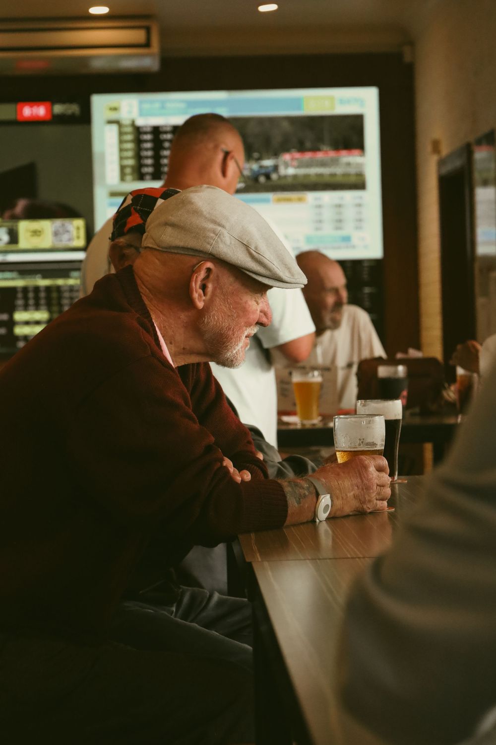 A person in a cap sits alone at a dimly lit bar counter with a glass of beer in front of them.