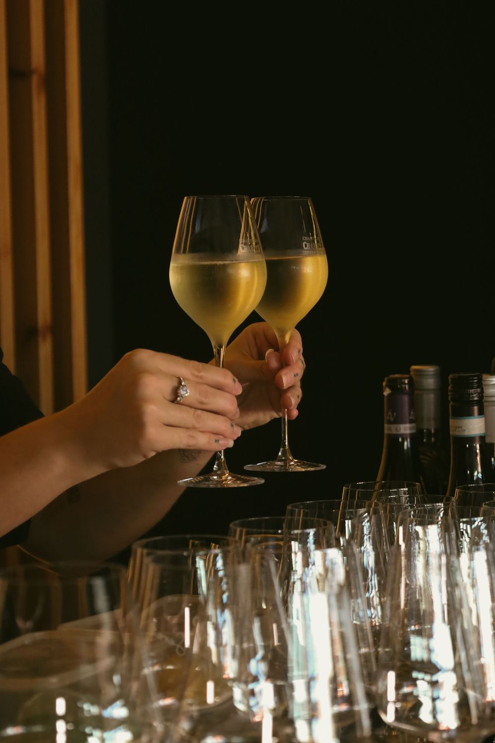 Two hands raise champagne glasses in a toast over a table filled with empty wine glasses in a dimly lit setting.