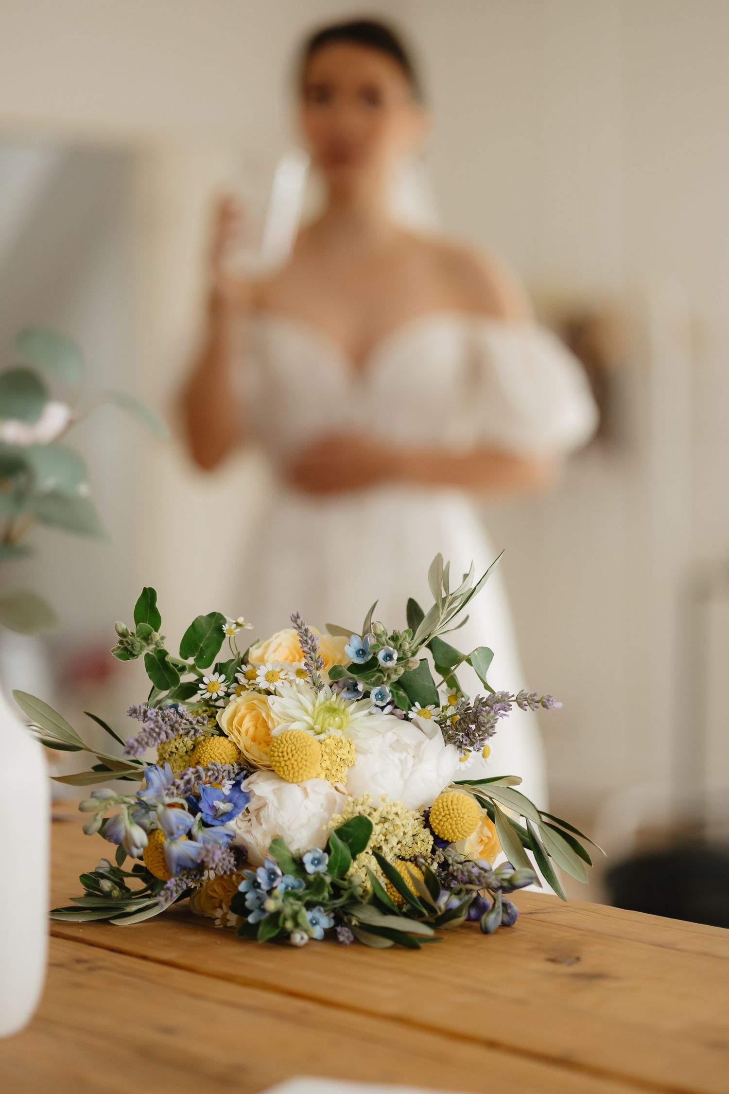 Delicate floral arrangement with yellow and blue flowers sits on wooden table with blurred background.