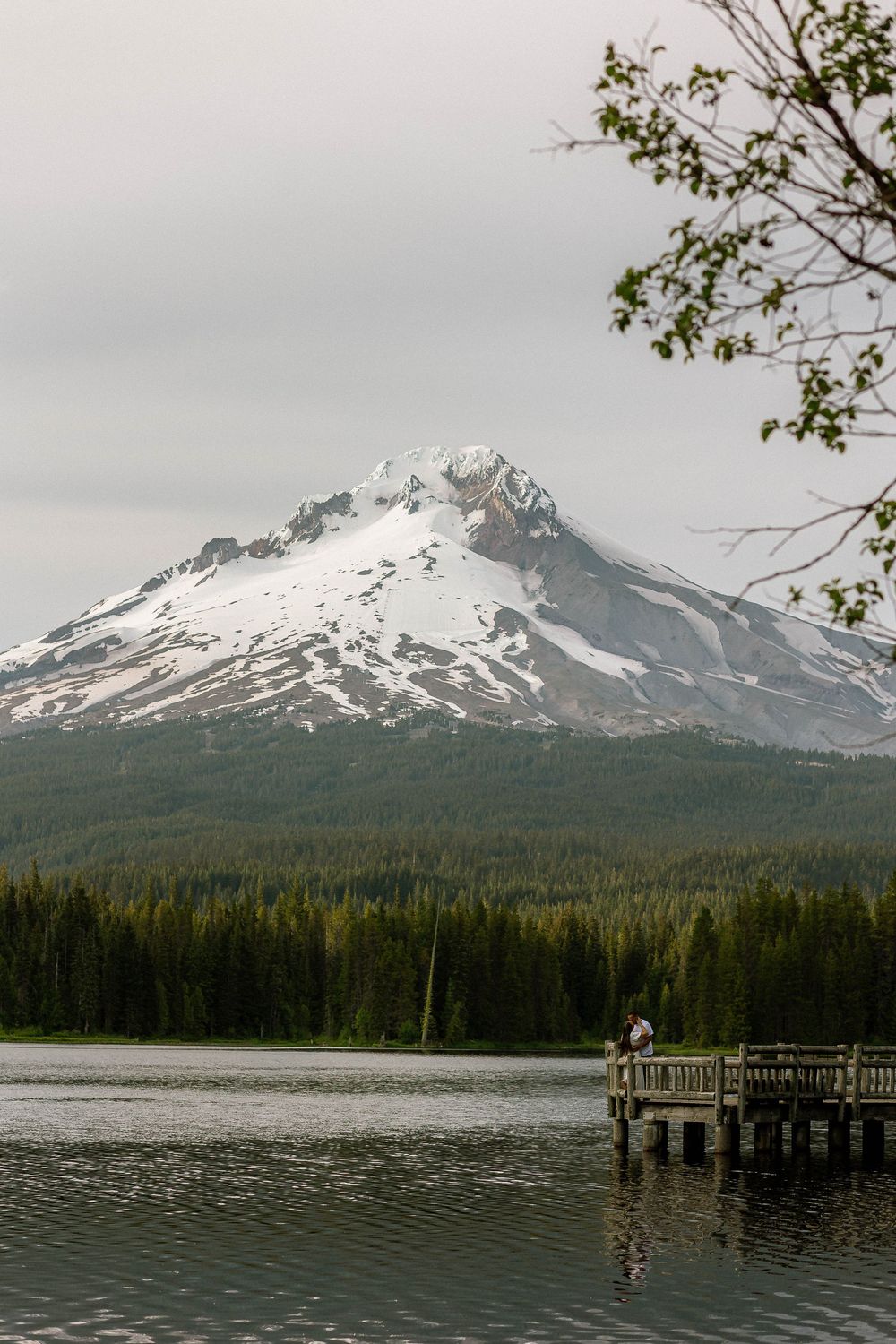 Person fishing on a dock with a snow-capped mountain and evergreen forest reflected in a calm lake.
