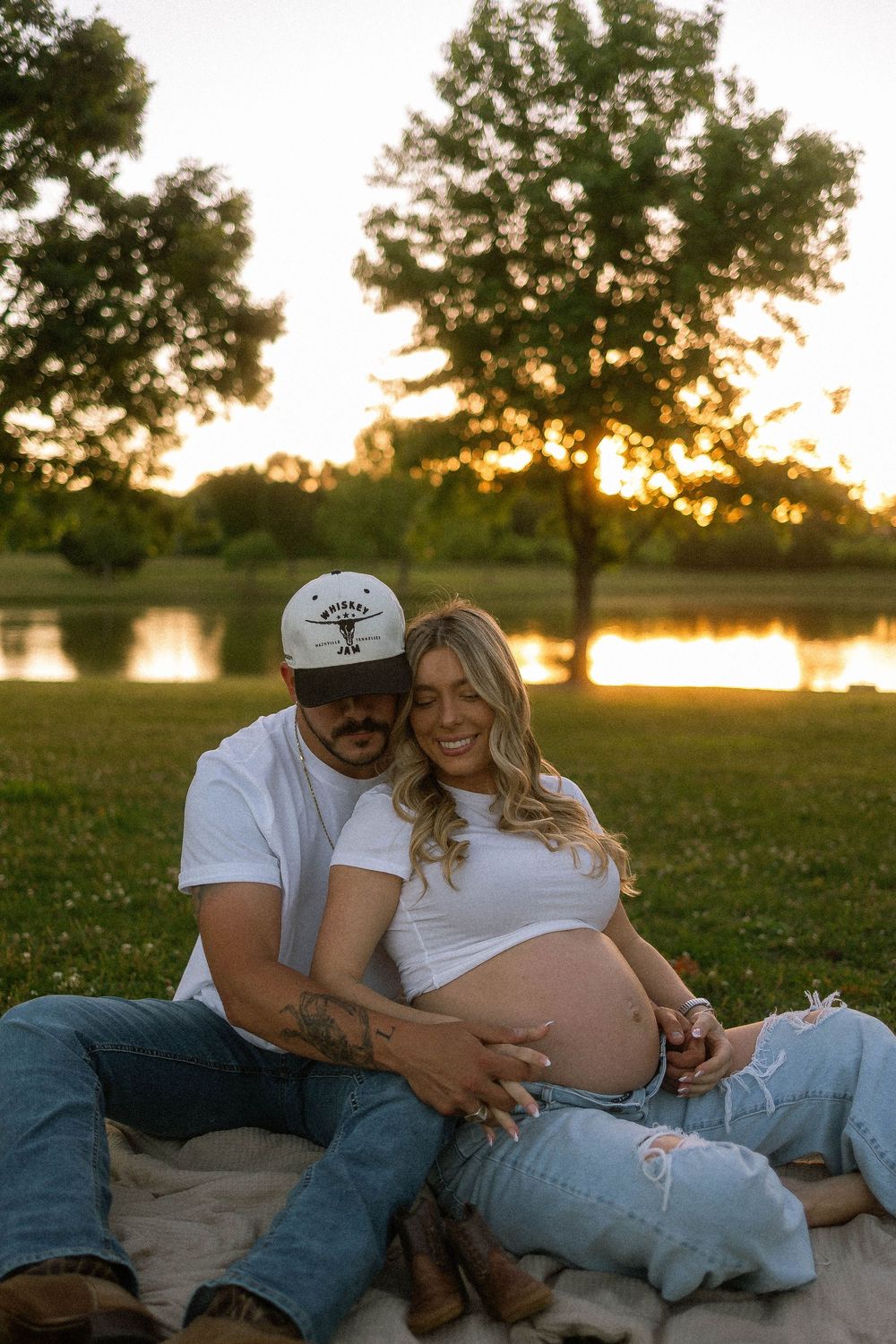 Couple sitting on blanket at sunset by lake, pregnant woman in white crop top with partner in cap cradling baby bump.