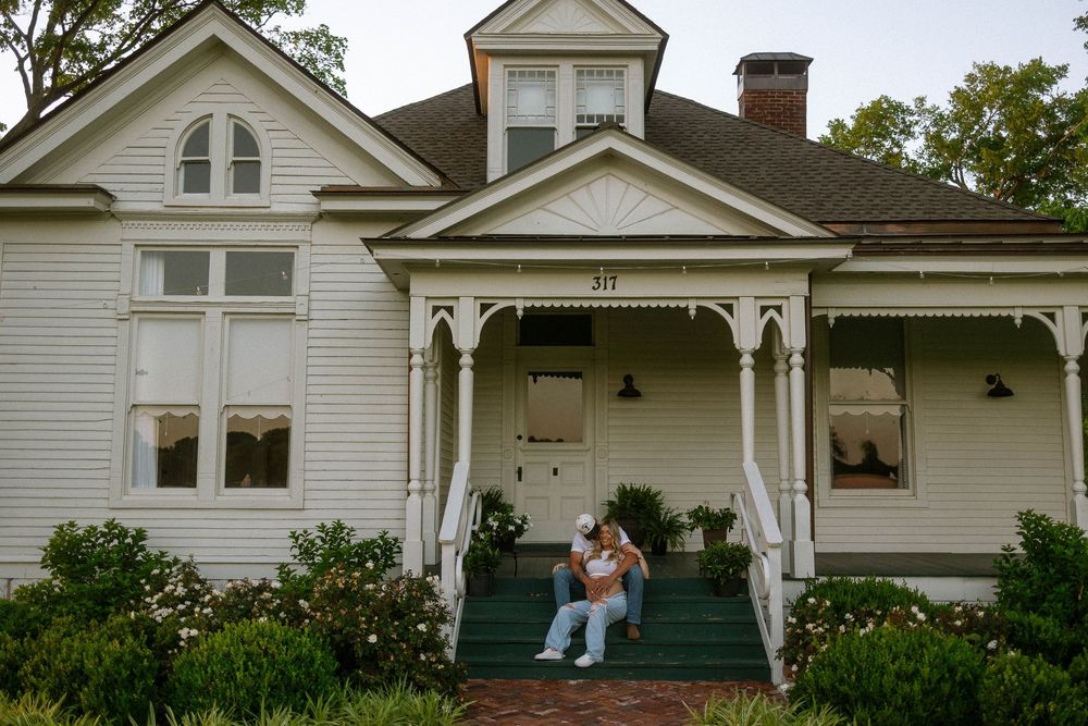 Couple sitting on porch steps of charming white Victorian home with decorative trim and lush green landscaping.