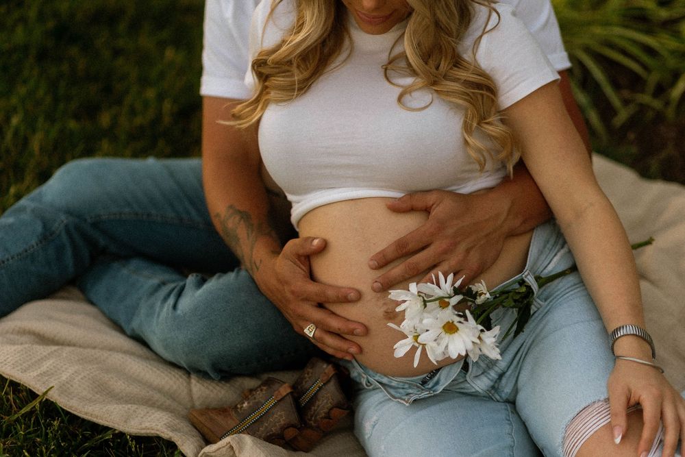 Pregnant woman in white crop top and jeans cradling baby bump outdoors with white daisies tucked in waistband.