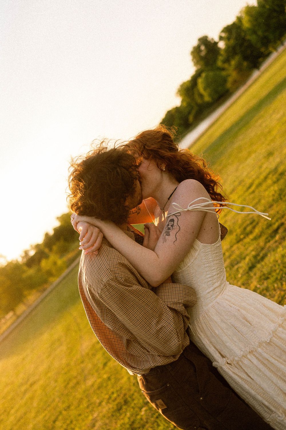Couple sharing a romantic kiss in a sunlit green field at golden hour, woman in white dress with tattoo.