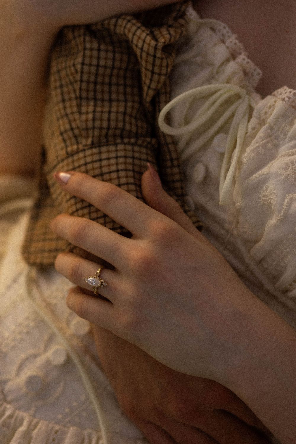 Close-up of a hand wearing a delicate gold marquise diamond ring, resting on vintage lace and plaid fabric.