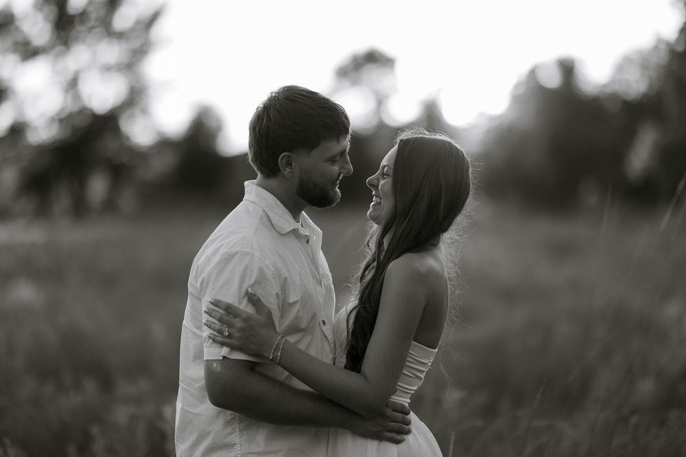 Black and white photo of a couple embracing and smiling at each other in a sunlit field.