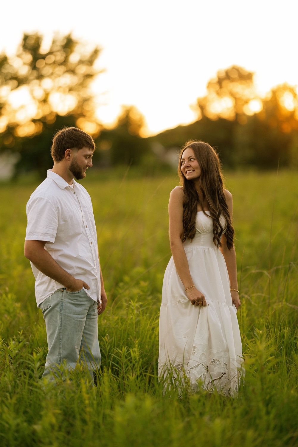Couple in white outfits standing in a lush green field at golden sunset, smiling at each other.