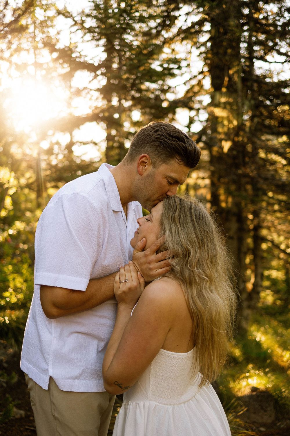 Couple sharing a tender kiss in golden sunlight among pine trees, dressed in white outfits for an engagement photo.